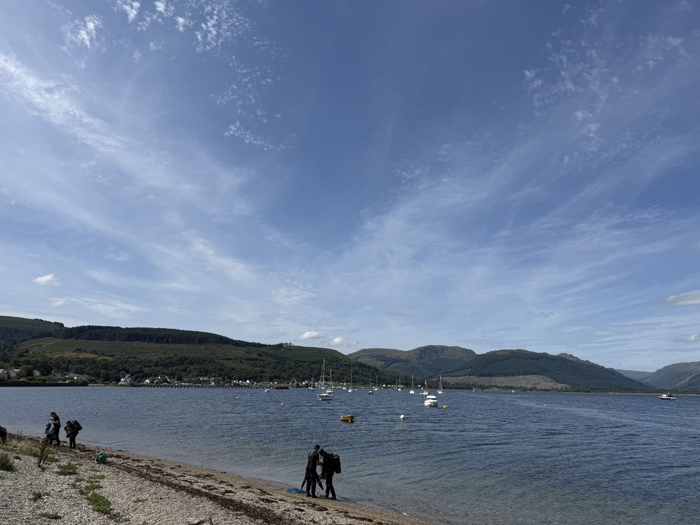 Boats on moorings off a shingle shore on a Scottish sea loch, with forested hills behind